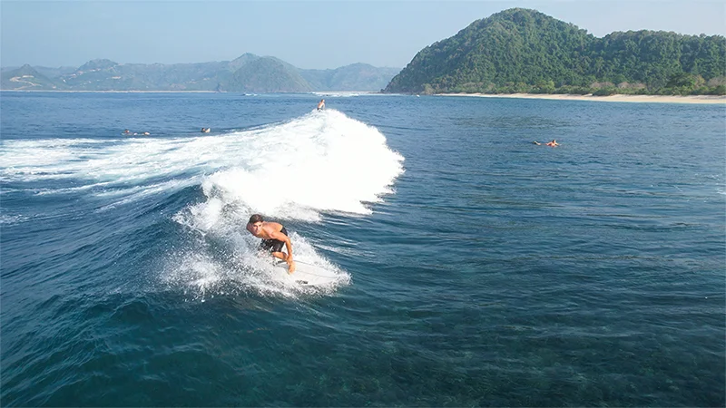 Surfer riding a wave at Mawi surf break in Lombok, Indonesia, with clear blue water and lush green hills in the background.
